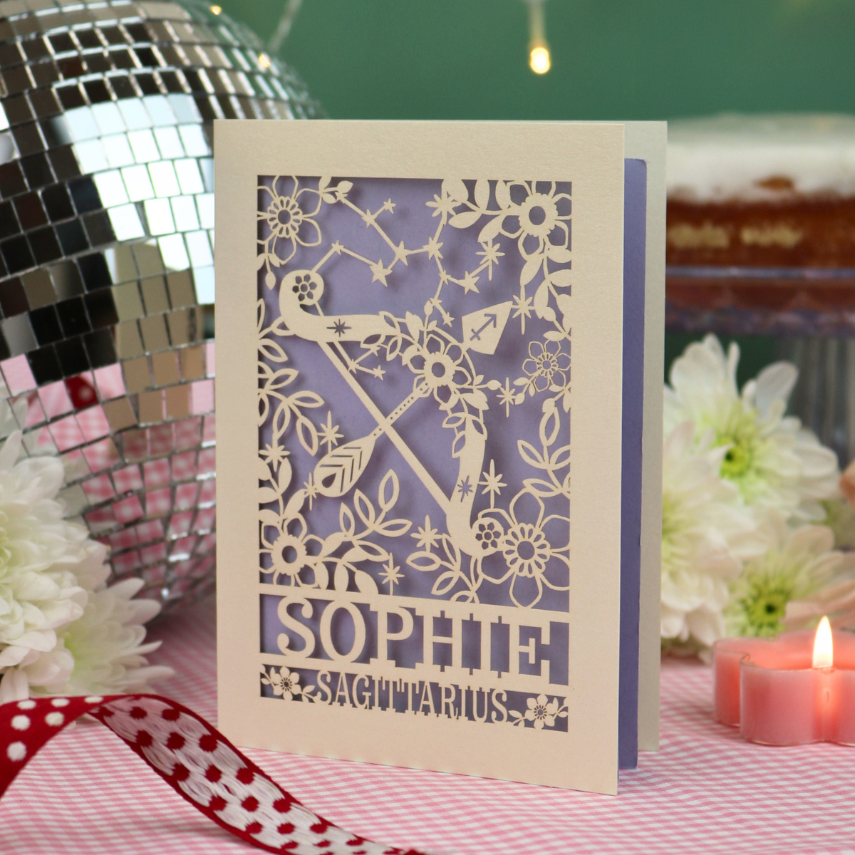 A laser-cut greeting card for the Sagittarius zodiac sign, featuring a cream-colored card with a lilac inner panel and cut out details of a bow and arrow, floral patterns, and the name 'SOPHIE'. The card is styled on a pink-and-white checked tablecloth next to a red polka-dot ribbon, a silver disco ball, and pink candles.