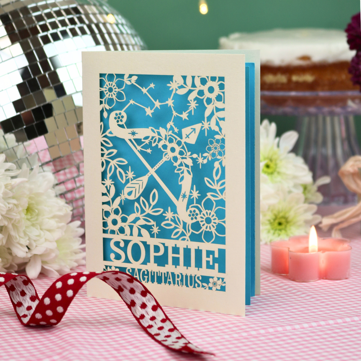 A personalized paper-cut birthday card with a blue and white Sagittarius design, standing on a pink checked tablecloth with a red polka-dot ribbon, white flowers, and a lit candle in the background.