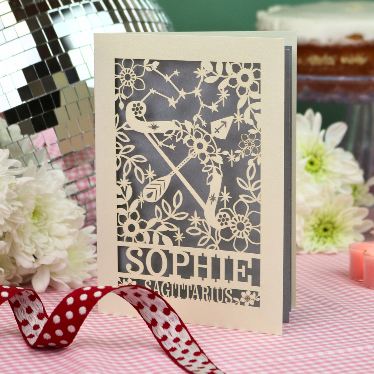 A personalized laser-cut greeting card for Sagittarius named 'Sophie' stands on a pink gingham tablecloth with a red and white polka dot ribbon in the foreground. The card features a cutout design of a bow and arrow surrounded by stars and flowers.