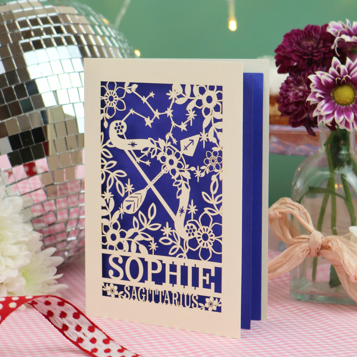 A handmade Sagittarius birthday card with a laser-cut design featuring a Sagittarius zodiac symbol, floral patterns, and the name 'SOPHIE' on a cream cardstock, styled on a pink checked tablecloth with a disco ball, flowers, and a ribbon.