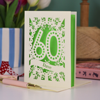 A paper cut out 60th birthday card, personalised with the name "Dave" in stencil lettering cut out from a banner shape. The card is laser cut from a metallic cream card and has a bright green paper insert. It stands on a white desk with flowers and stationery items in the background.