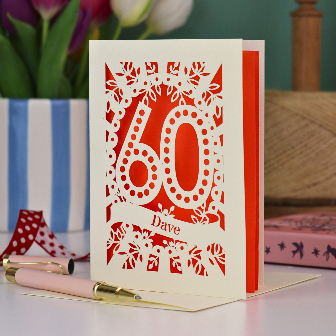 A paper cut out 60th birthday card, personalised with the name "Dave" in stencil lettering cut out from a banner shape. The card is laser cut from a metallic cream card and has a bright orange paper insert. It stands on a white desk with flowers and stationery items in the background.
