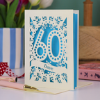 A paper cut out 60th birthday card, personalised with the name "Dave" in stencil lettering cut out from a banner shape. The card is laser cut from a metallic cream card and has a bright blue paper insert. It stands on a white desk with flowers and stationery items in the background.