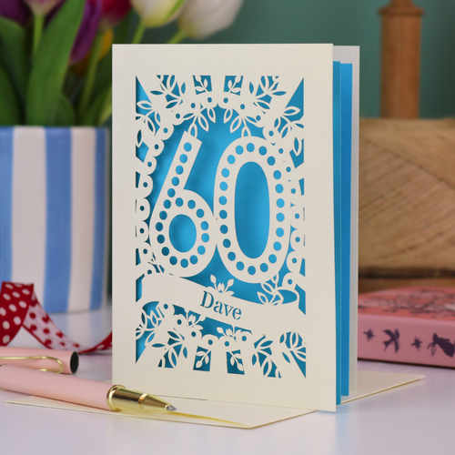 A paper cut out 60th birthday card, personalised with the name "Dave" in stencil lettering cut out from a banner shape. The card is laser cut from a metallic cream card and has a bright blue paper insert. It stands on a white desk with flowers and stationery items in the background.