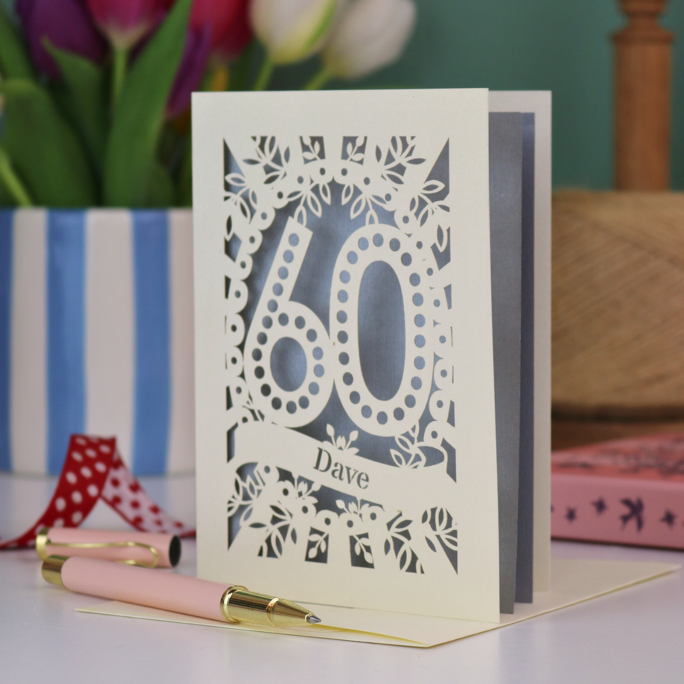 A paper cut out 60th birthday card, personalised with the name "Dave" in stencil lettering cut out from a banner shape. The card is laser cut from a metallic cream card and has a silver paper insert. It stands on a white desk with flowers and stationery items in the background.