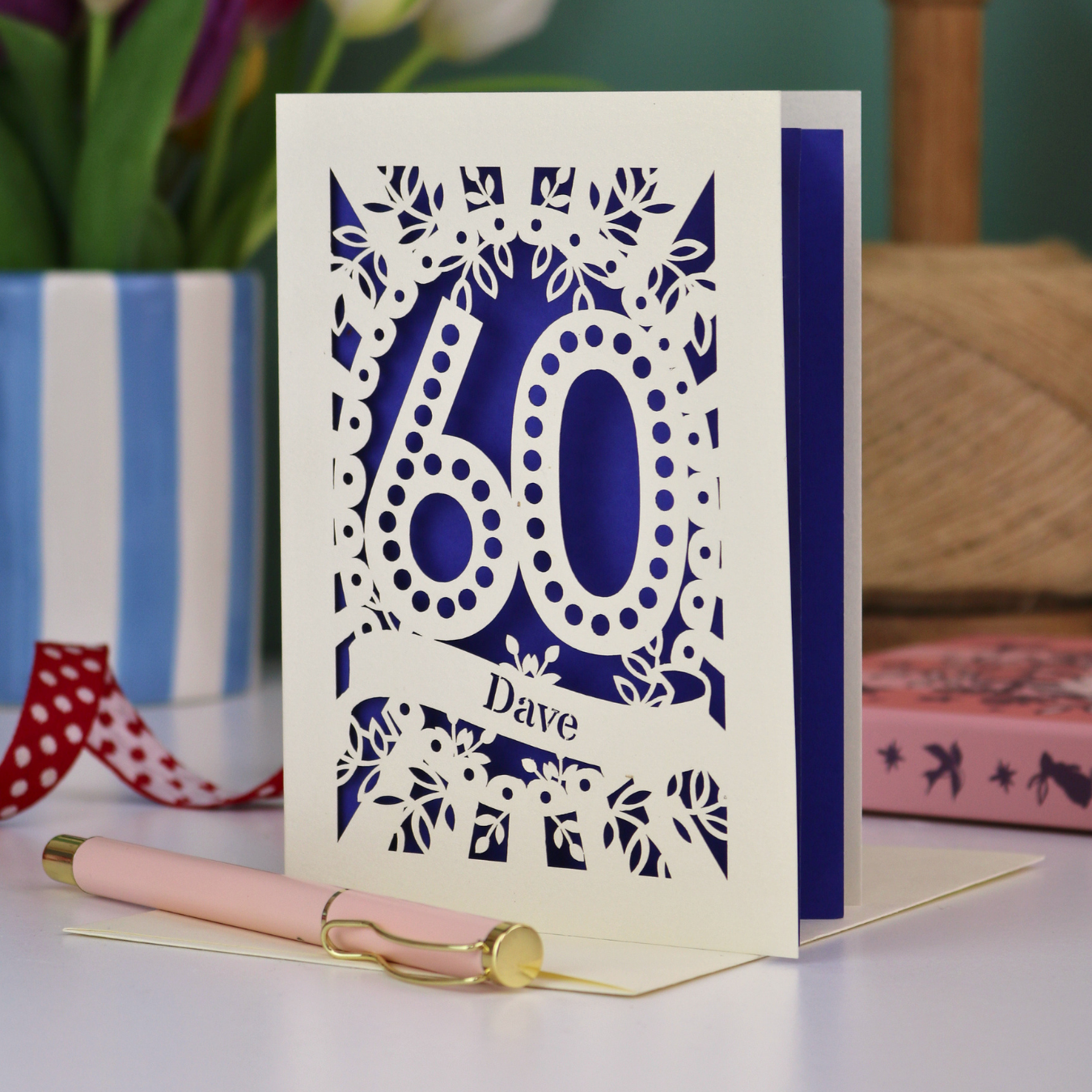 A paper cut out 60th birthday card, personalised with the name "Dave" in stencil lettering cut out from a banner shape. The card is laser cut from a metallic cream card and has a violet paper insert. It stands on a white desk with flowers and stationery items in the background.