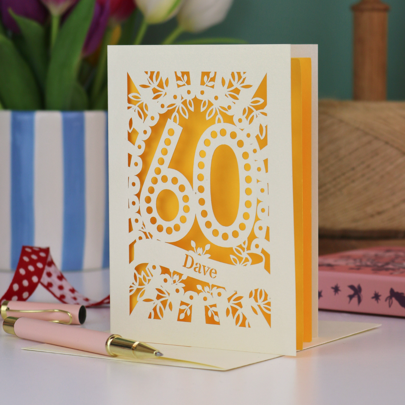 A paper cut out 60th birthday card, personalised with the name "Dave" in stencil lettering cut out from a banner shape. The card is laser cut from a metallic cream card and has a yellow paper insert. It stands on a white desk with flowers and stationery items in the background.
