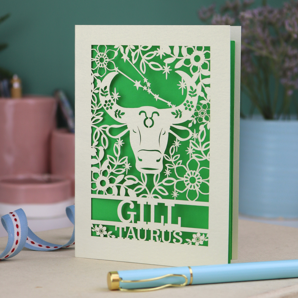 Personalised laser cut Birthday card with zodiac design featuring a bull on a pink background, with a pen and decorative ribbon in the foreground.