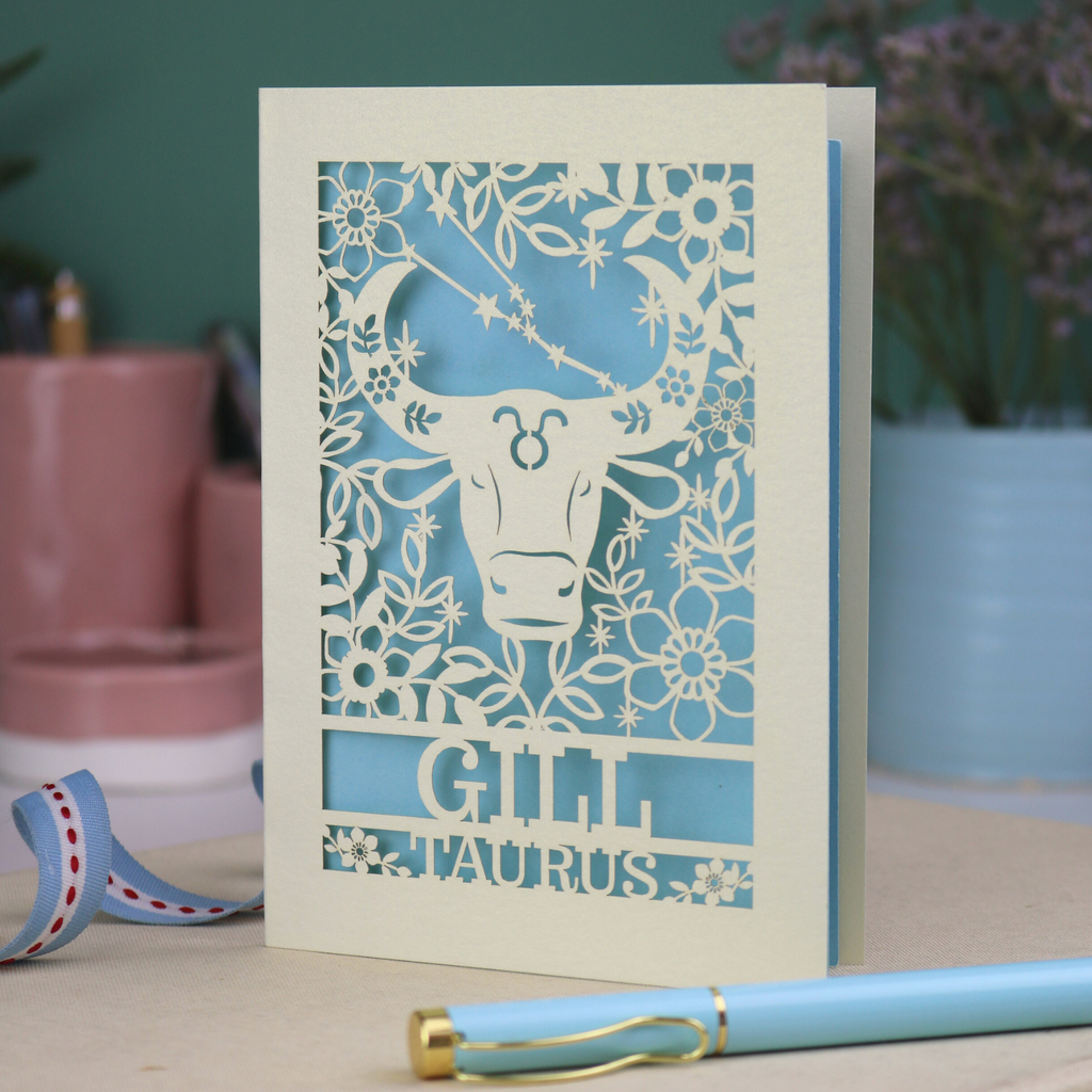 Personalised laser cut Birthday card with zodiac design featuring a bull on a pink background, with a pen and decorative ribbon in the foreground.