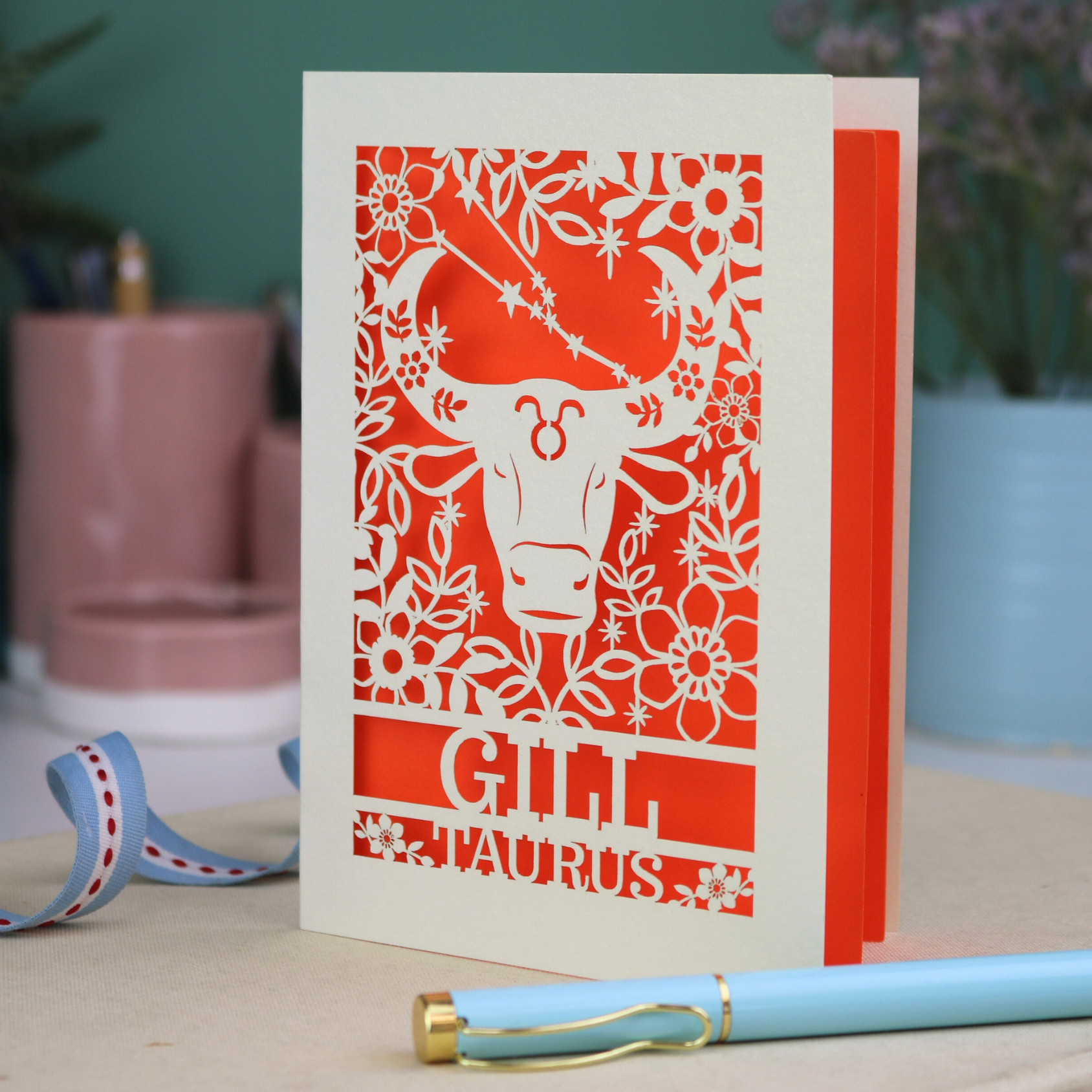 Personalised laser cut Birthday card with zodiac design featuring a bull on a pink background, with a pen and decorative ribbon in the foreground.