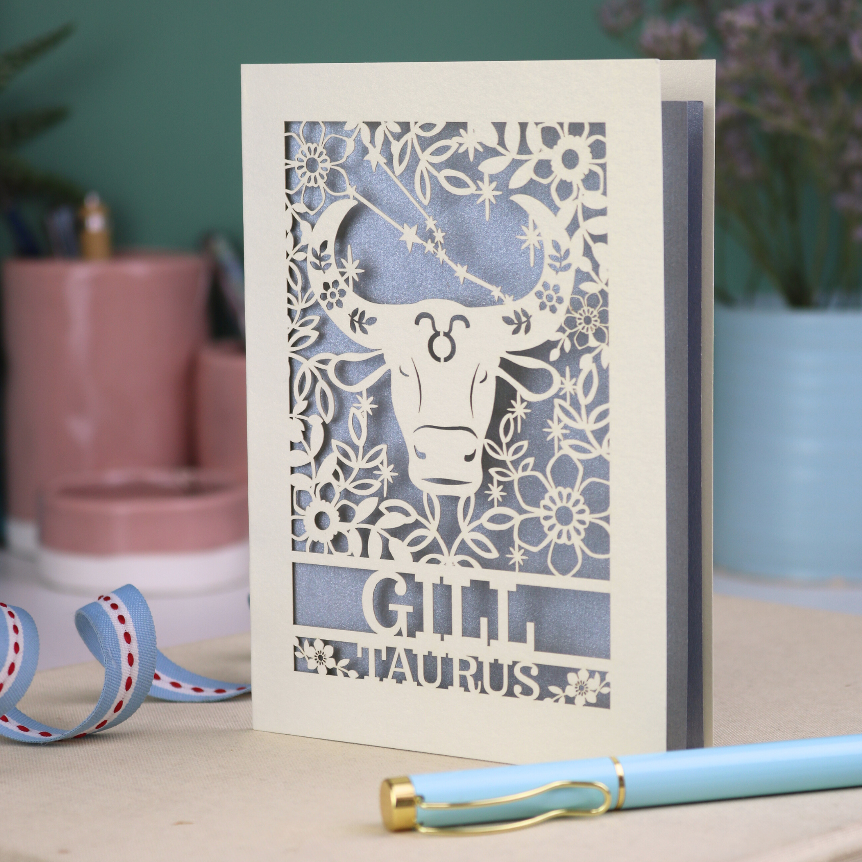 Personalised laser cut Birthday card with zodiac design featuring a bull on a pink background, with a pen and decorative ribbon in the foreground.