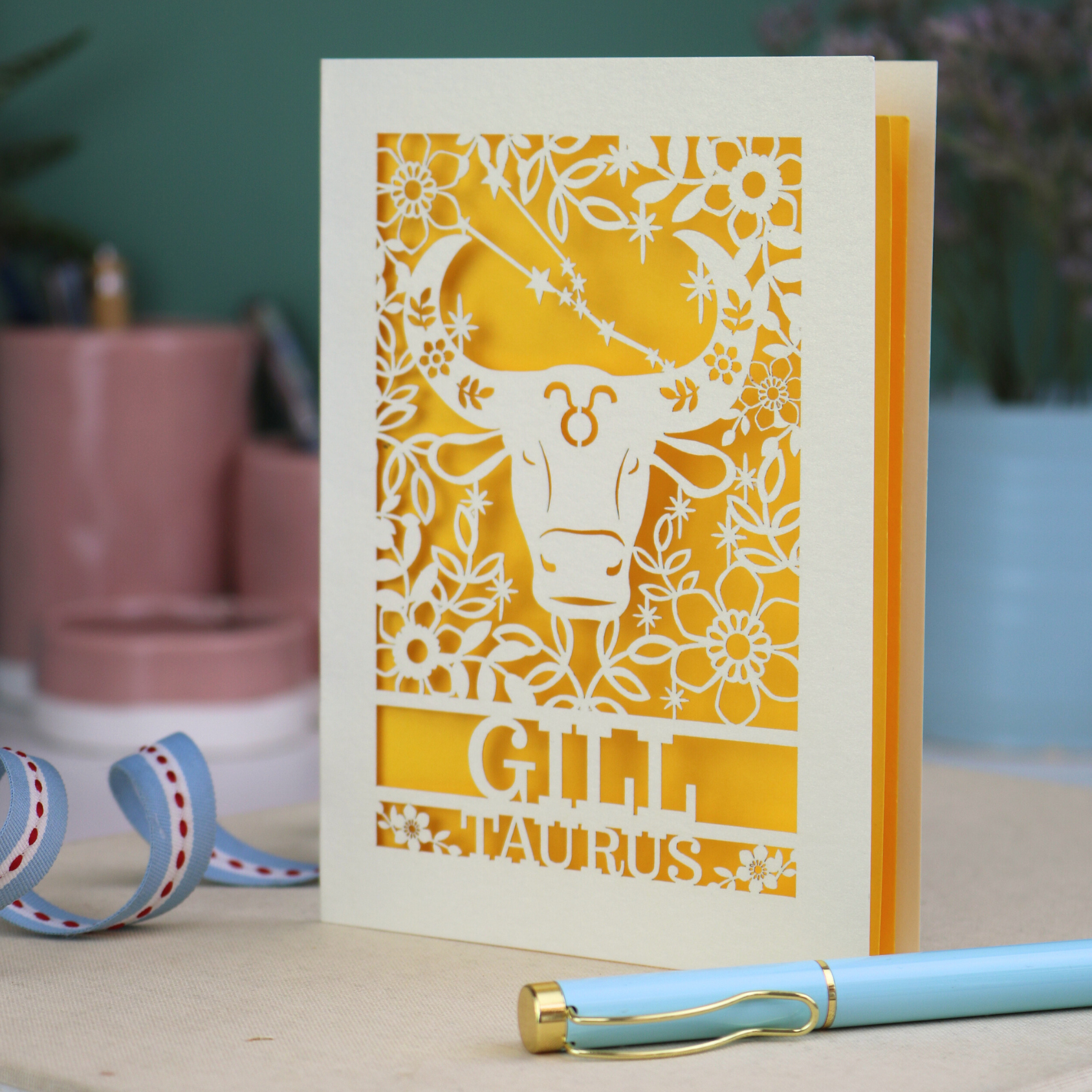 Personalised laser cut Birthday card with zodiac design featuring a bull on a pink background, with a pen and decorative ribbon in the foreground.