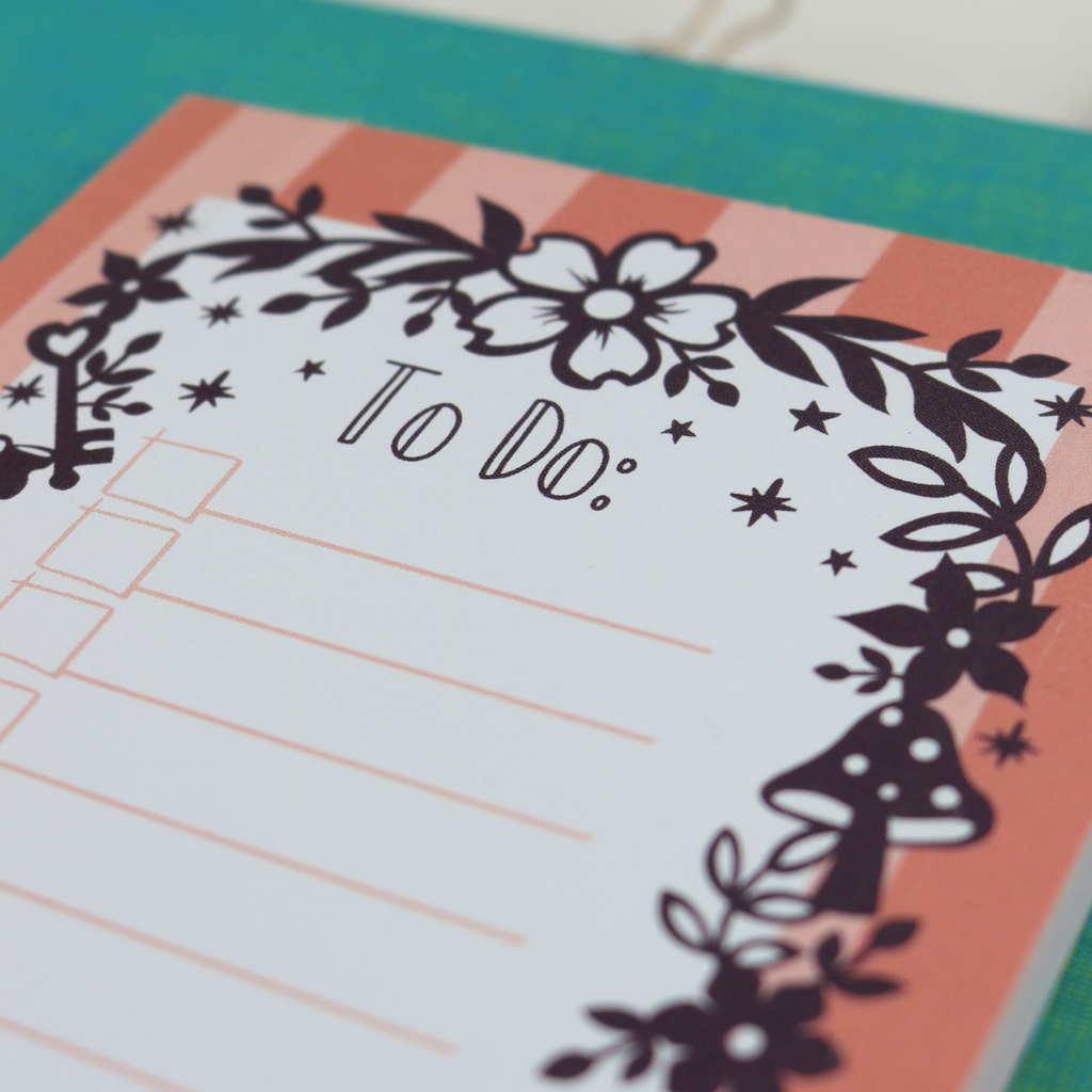 A close up detail of a notepad with pink striped border and purple floral illustrations, with "To Do:" at the top and lines with check boxes