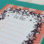 A close up detail of a notepad with pink striped border and purple floral illustrations, with "To Do:" at the top and lines with check boxes
