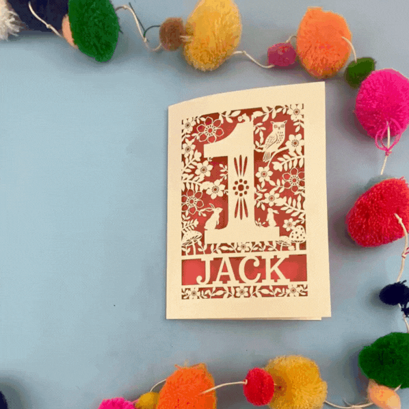 Personalised Laser cut First Birthday Card with 'Jack' design surrounded by colourful pom-poms on a blue background