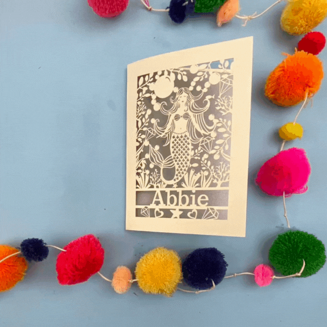 Paper cut out birthday card opening and closing in stop motion, with a mermaid and 'Abbie' on a blue background surrounded by colourful pom-poms.