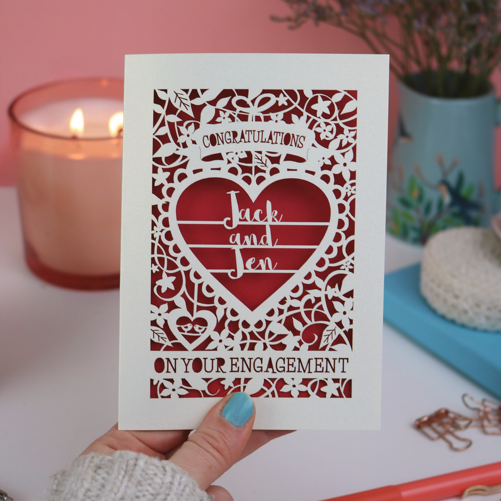 A cream laser cut engagement card on a desk with stationery items. The card is portrait-orientation, with floral cut out details and 'Congratulations, name and name, on your engagement'