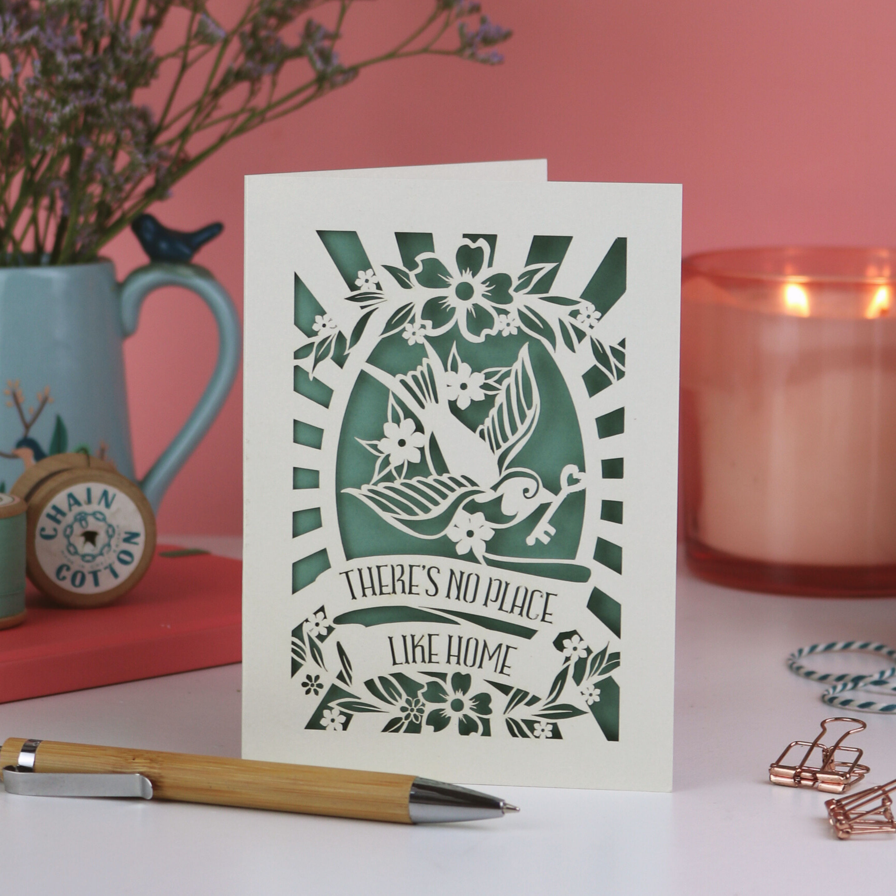 Decorative laser cut new home card with green cut-out design and text, placed on a surface with a pen, mug, and candle in the background.