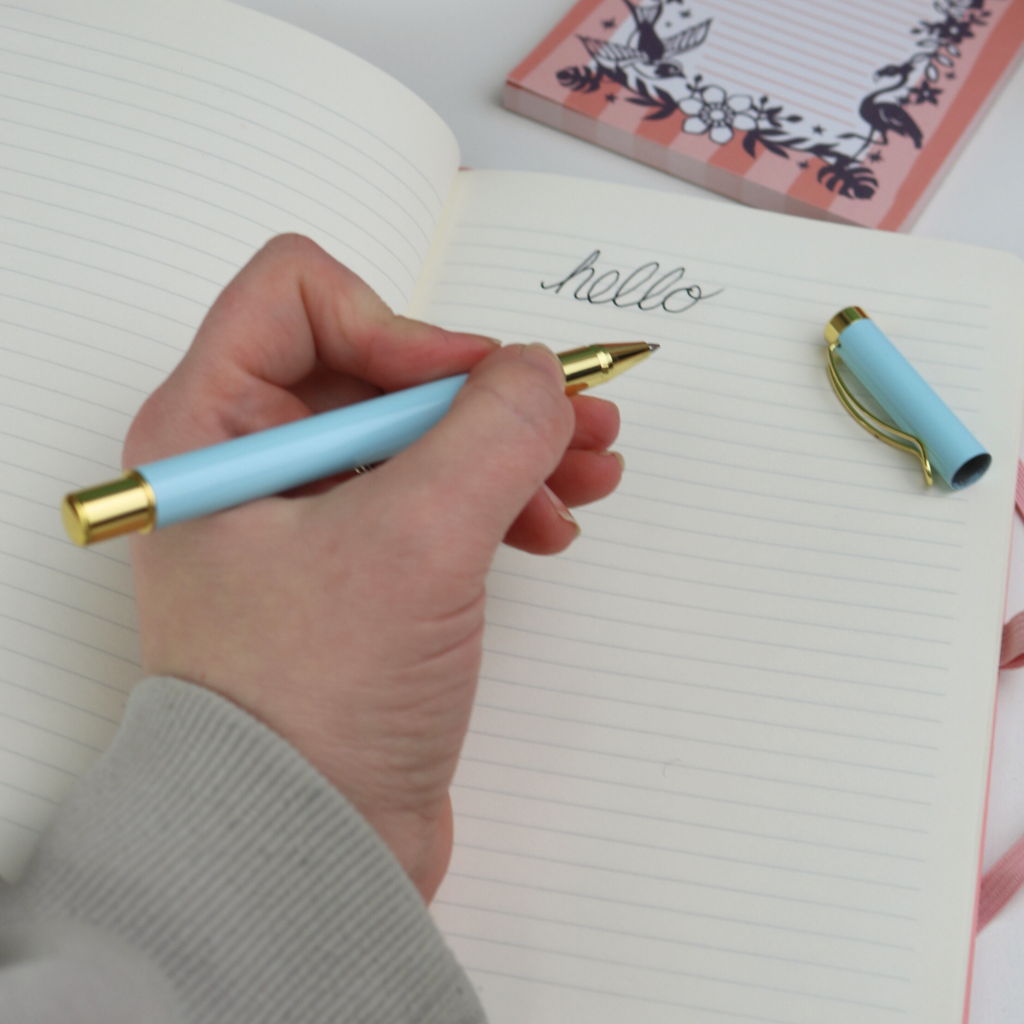 Hand holding a light blue pen with gold accents, writing 'hello' on a notebook.