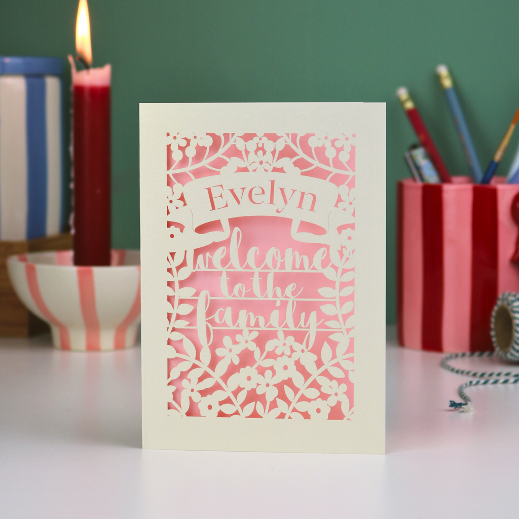 Decorative card with 'Evelyn welcome to the family' text on a pink background, placed on a surface with candles and stationery items in the background.