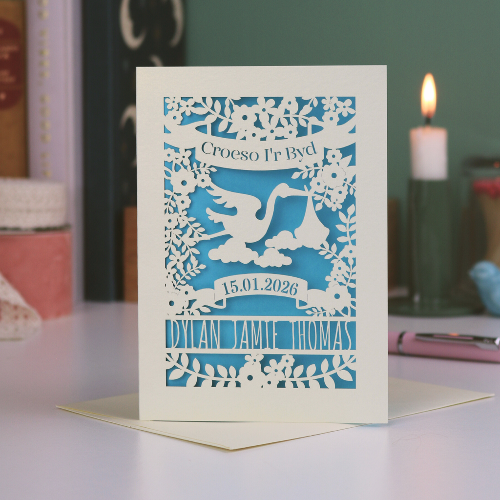A laser cut new baby card with Welsh text, "Croeso I'R Byd", a stork and floral cut out design and the baby's date of birth and name. 
