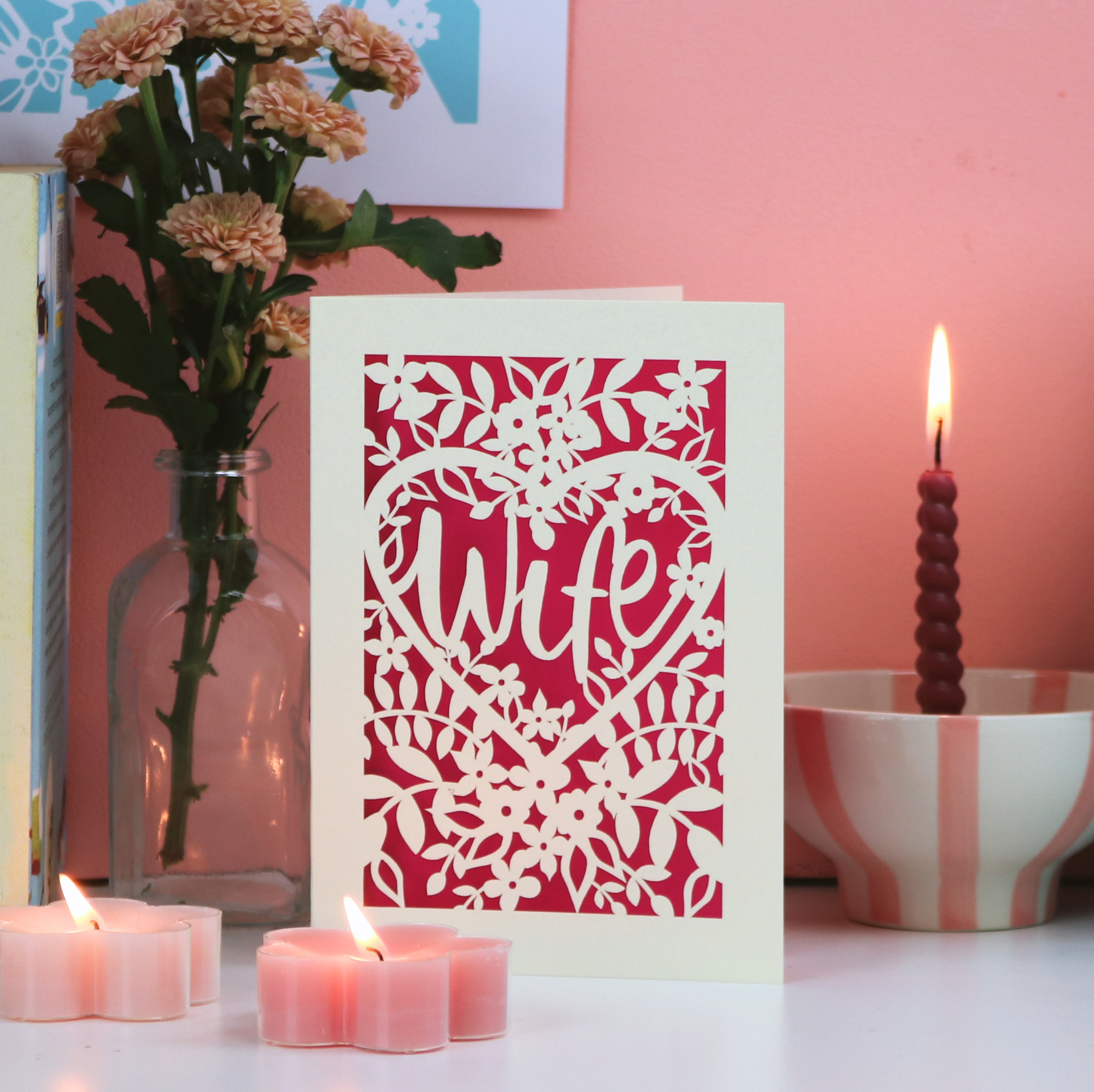 A laser cut floral Valentine's card that has the word "Wife" in a heart surrounded by a floral cut out design, on a white desk with candles and flowers, with a pink background