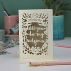 Laser-cut card asking 'Will you be my bridesmaid?' on a desk with stationery items.