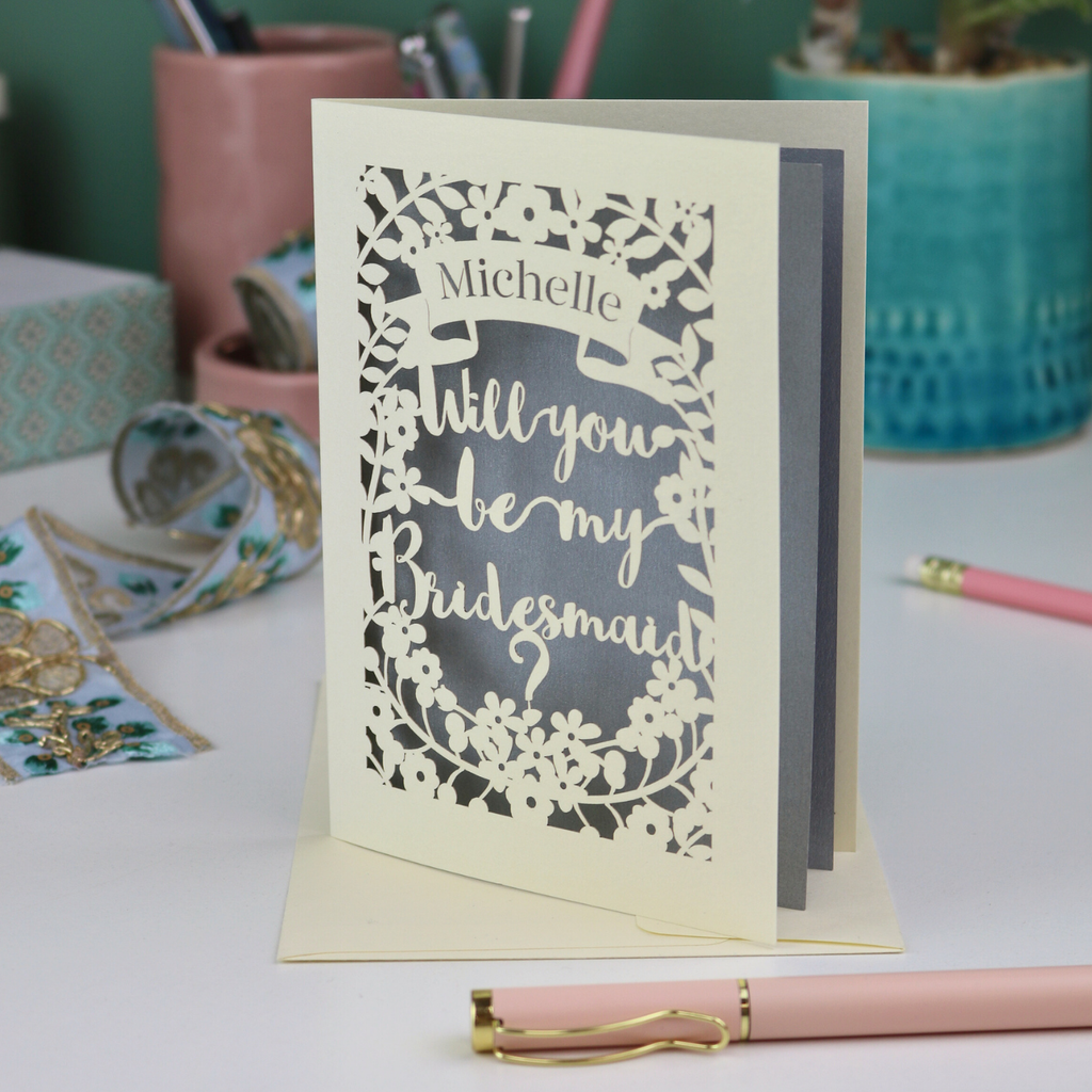 Laser-cut card asking 'Will you be my bridesmaid?' on a desk with stationery items.