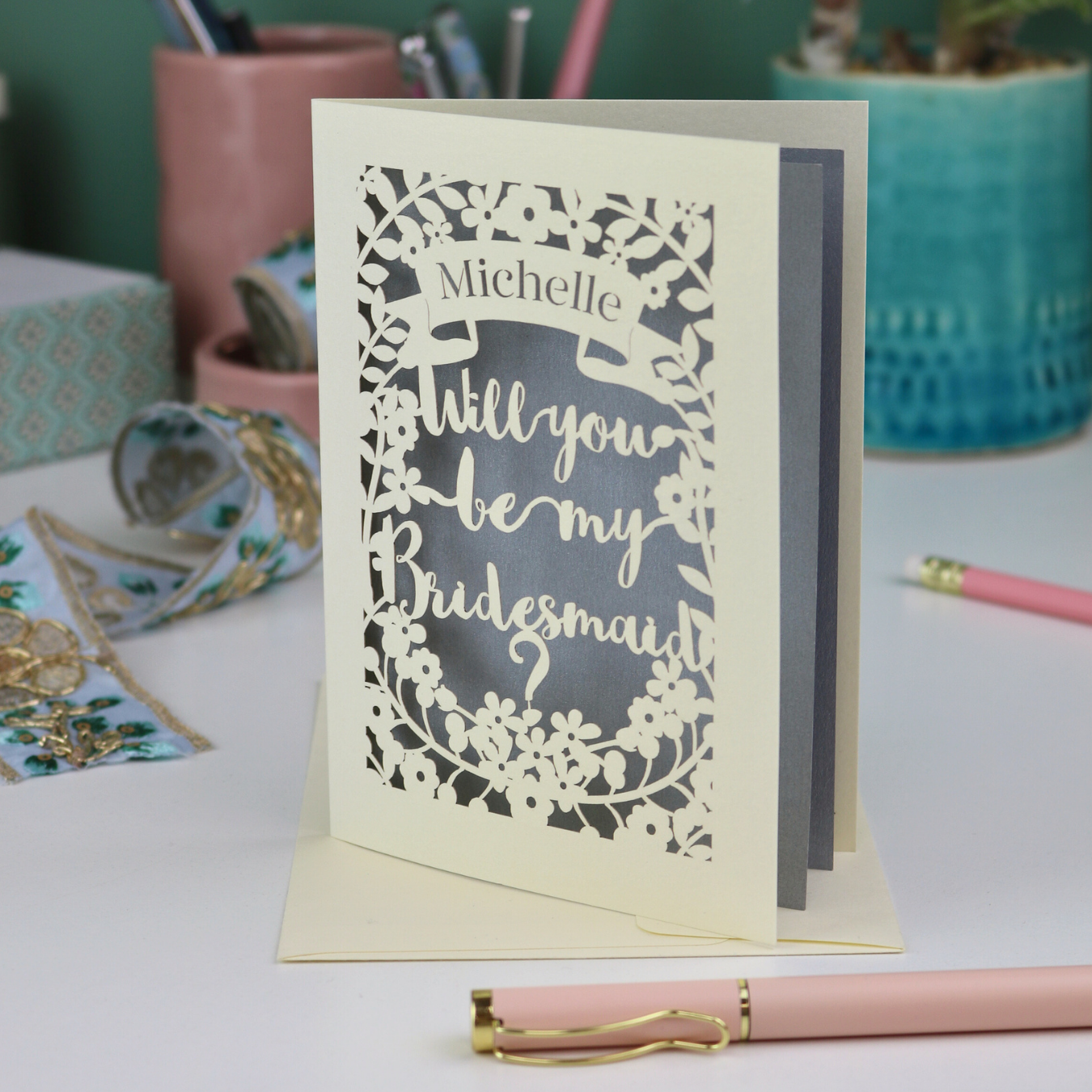 Laser-cut card asking 'Will you be my bridesmaid?' on a desk with stationery items.
