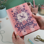 Hands holding a pink A5 notebook with purple illustrations and text "All of my Best Ideas"