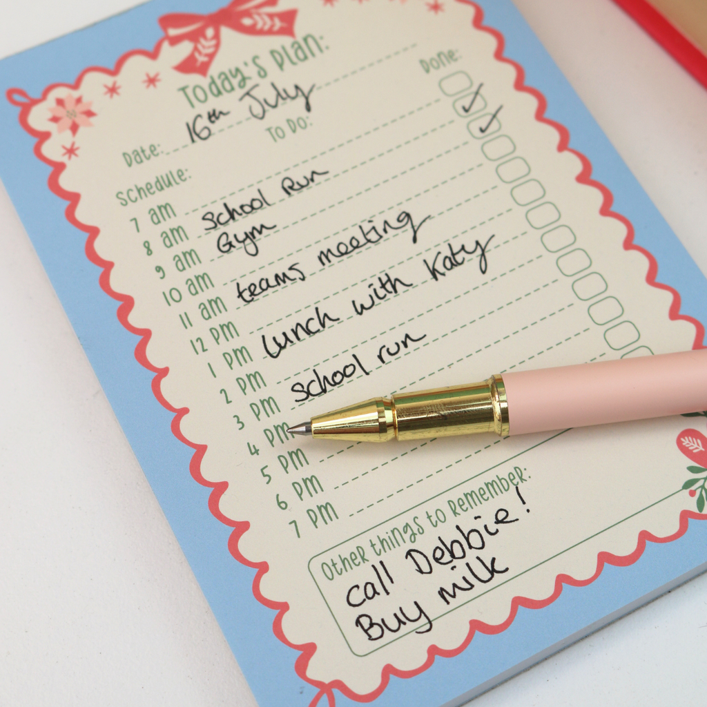 A close-up of a pink and gold pen resting on a decorated daily planner pad. The planner has a light blue border with a red scalloped inner frame and handwritten notes, photographed on a white surface.