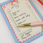 A close-up of a pink and gold pen resting on a decorated daily planner pad. The planner has a light blue border with a red scalloped inner frame and handwritten notes, photographed on a white surface.