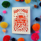 Adoption Card with 'Robyn Welcome to the Family' text on a blue background with colourful pom-poms.