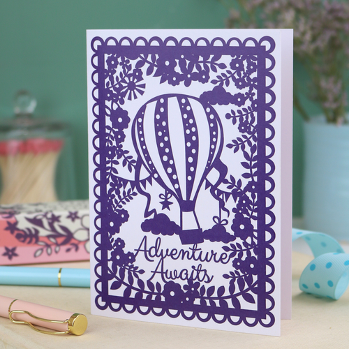 Printed card with floral purple border and hot air balloon design with "Adventure awaits" written in a cursive font