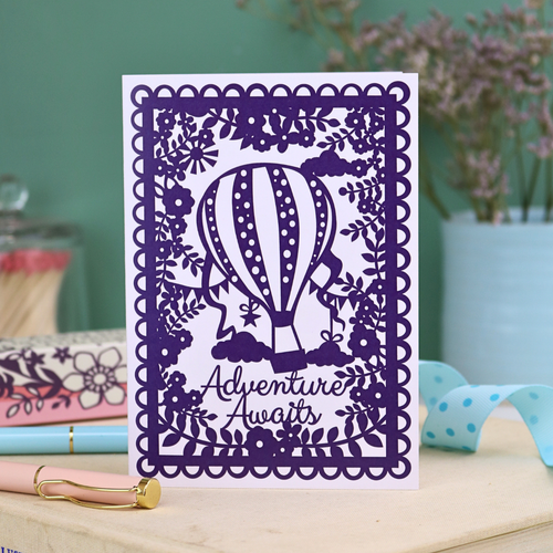 Printed graduation card with floral purple border and hot air balloon design with "Adventure awaits" written in a cursive font