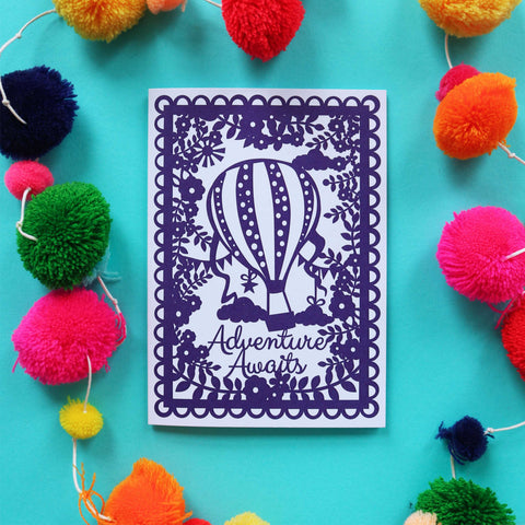 A printed card that looks like a papercut, with a hot air balloon and the words "Adventure Awaits"
