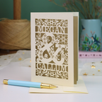 A personalized laser-cut anniversary card with floral patterns and the names 'MEGAN & CALLUM', displayed on a table with a matching light blue and gold pen in front.