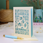 A personalized laser-cut anniversary card with floral patterns and the names 'MEGAN & CALLUM', displayed on a table with a matching light blue and gold pen in front.
