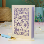A personalized cut out anniversary card with floral patterns and the names 'MEGAN & CALLUM', displayed on a table with a matching light blue and gold pen in front.
