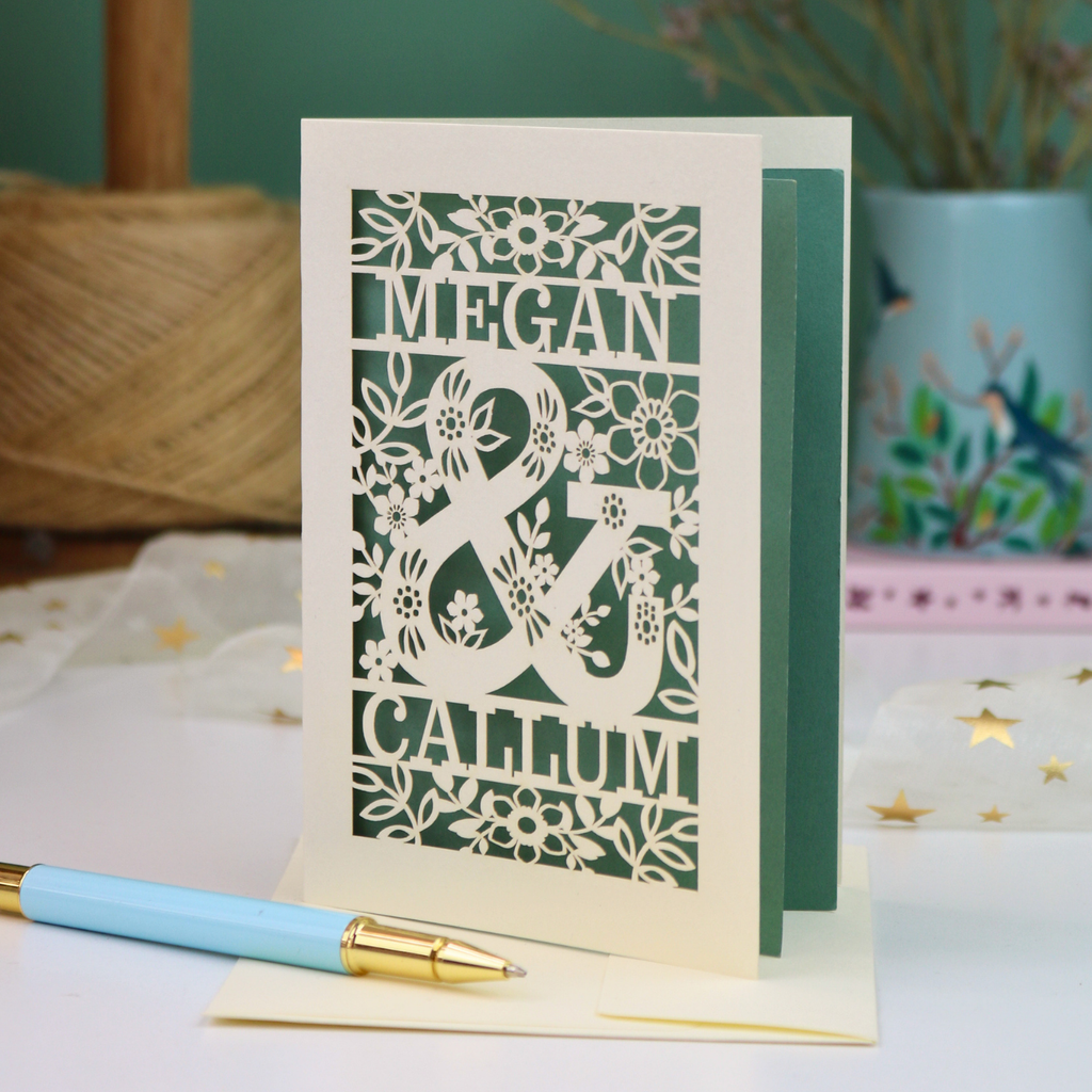 A personalised laser-cut anniversary card with floral patterns and the names 'MEGAN & CALLUM', displayed on a table with a matching light blue and gold pen in front.