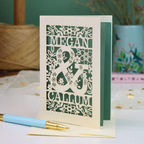 A personalised laser-cut anniversary card with floral patterns and the names 'MEGAN & CALLUM', displayed on a table with a matching light blue and gold pen in front.