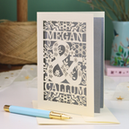 A laser-cut wedding anniversary card with floral patterns and the names 'MEGAN & CALLUM' in cut out lettering, displayed on a table with a light blue and gold pen in front.