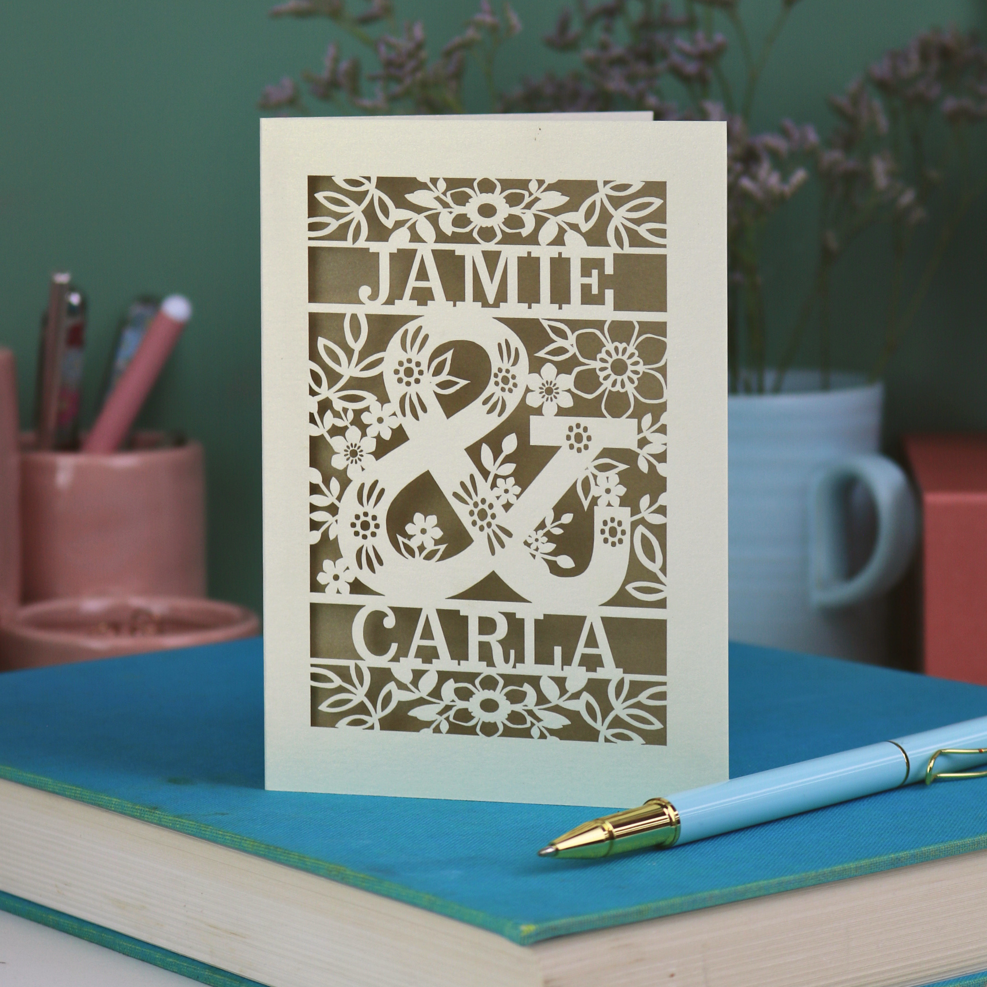 Personalised Engagement Card with laser-cut design of 'Jamie & Carla' on a table with stationery items.
