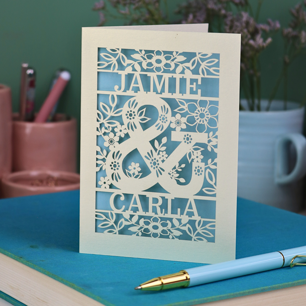 Personalised Engagement Card with laser-cut design of 'Jamie & Carla' on a table with stationery items.