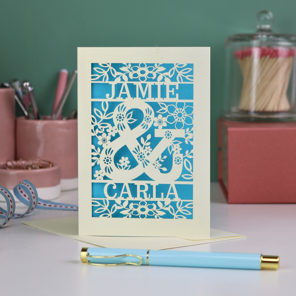 Personalised Engagement Card with laser-cut design of 'Jamie & Carla' on a table with stationery items.