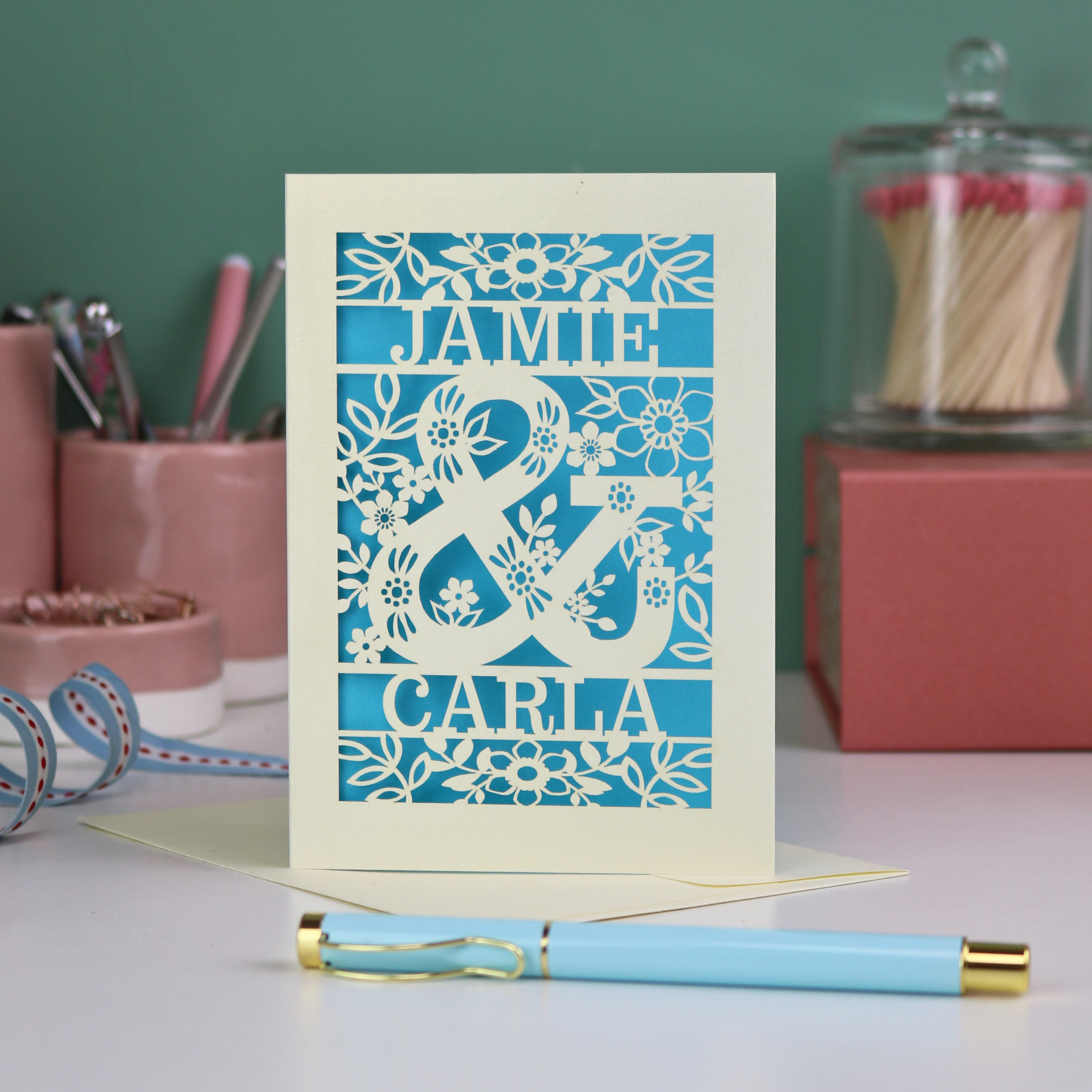 Personalised Engagement Card with laser-cut design of 'Jamie & Carla' on a table with stationery items.