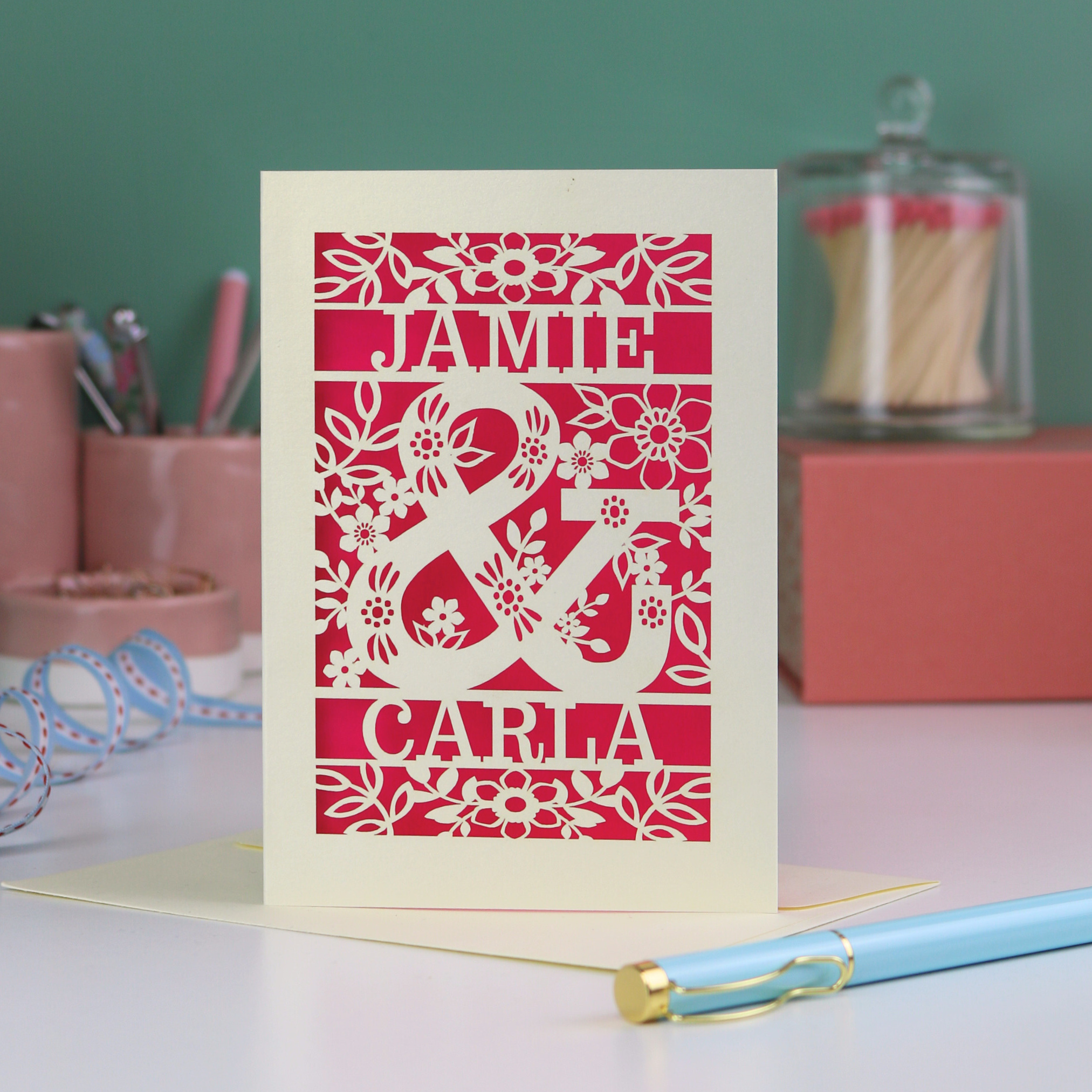 Personalised Engagement Card with laser-cut design of 'Jamie & Carla' on a table with stationery items.