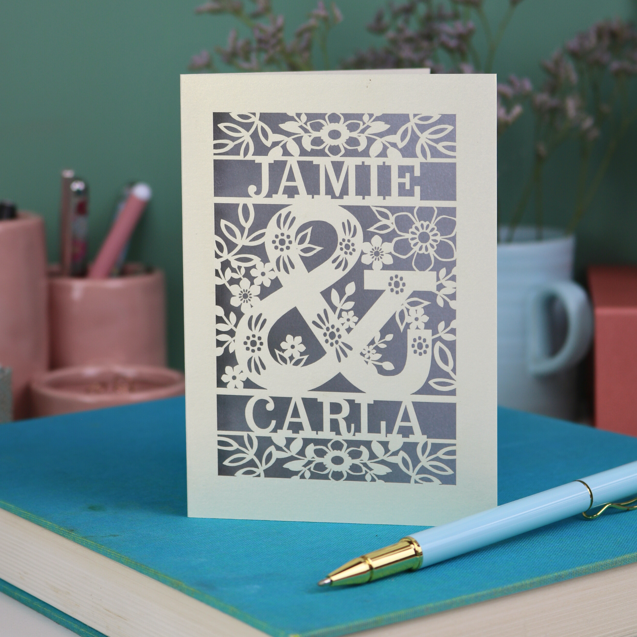 Personalised Engagement Card with laser-cut design of 'Jamie & Carla' on a table with stationery items.