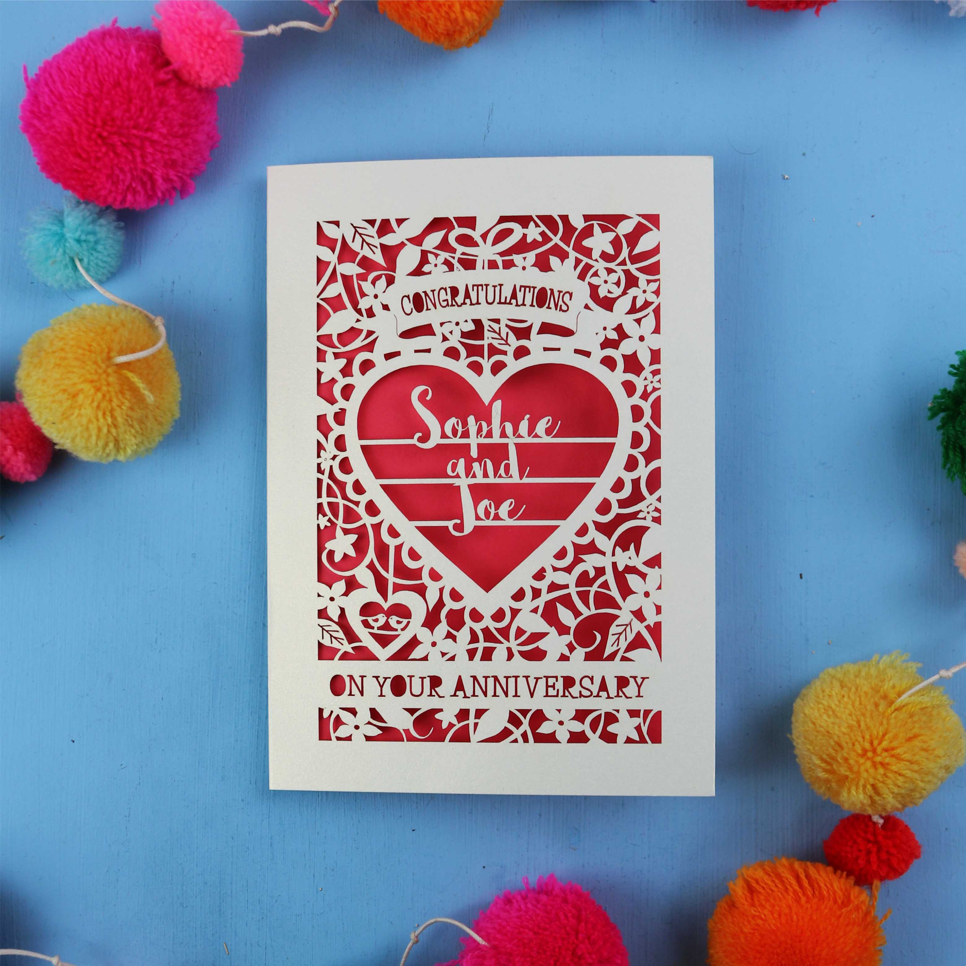 Cut out Anniversary card with red heart design on a blue background with colourful pom-poms