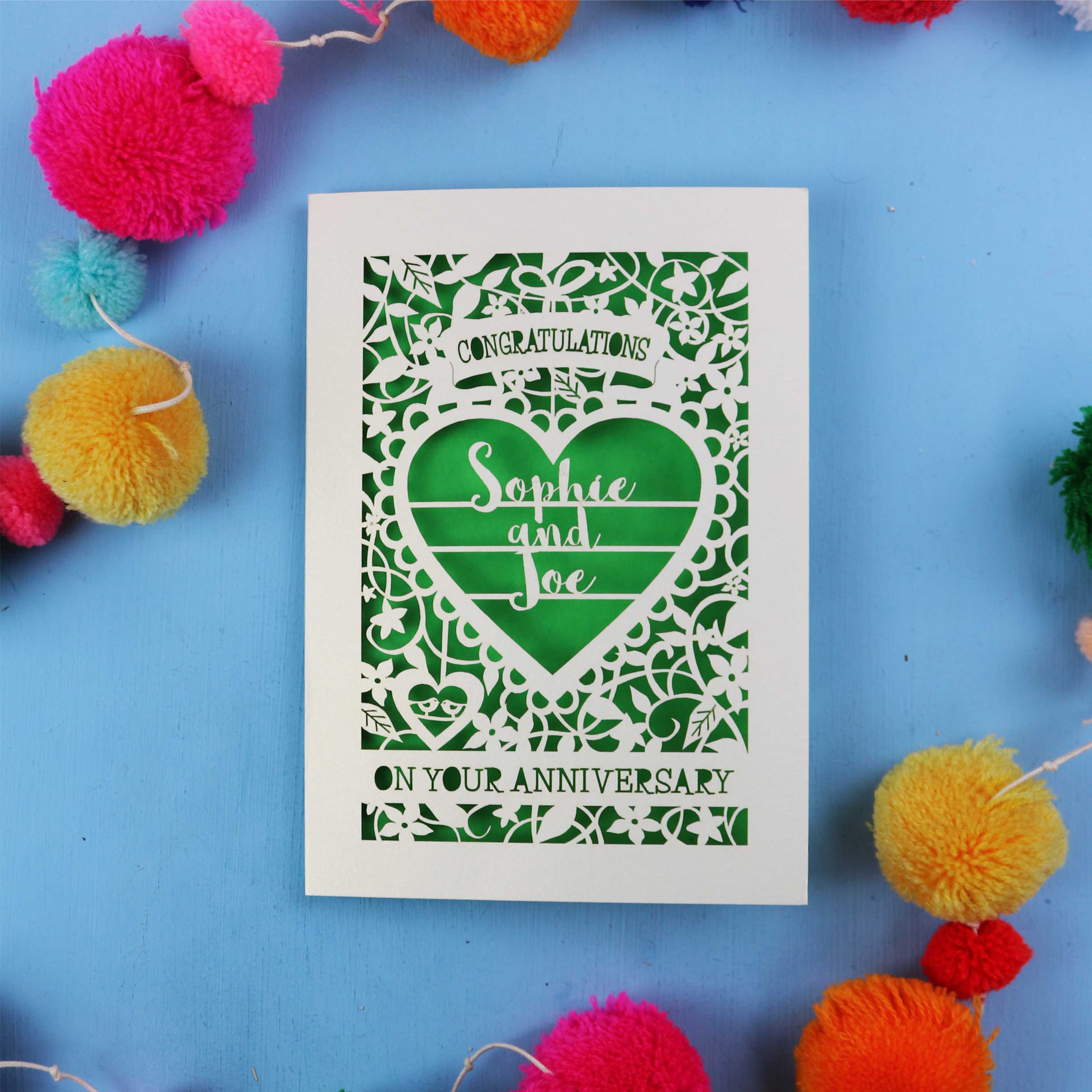 Personalised emerald wedding anniversary card with decorative heart design on a blue background with colourful pom-poms.